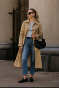 Woman wearing a beige trench coat, blue jeans, and black loafers standing outdoors.