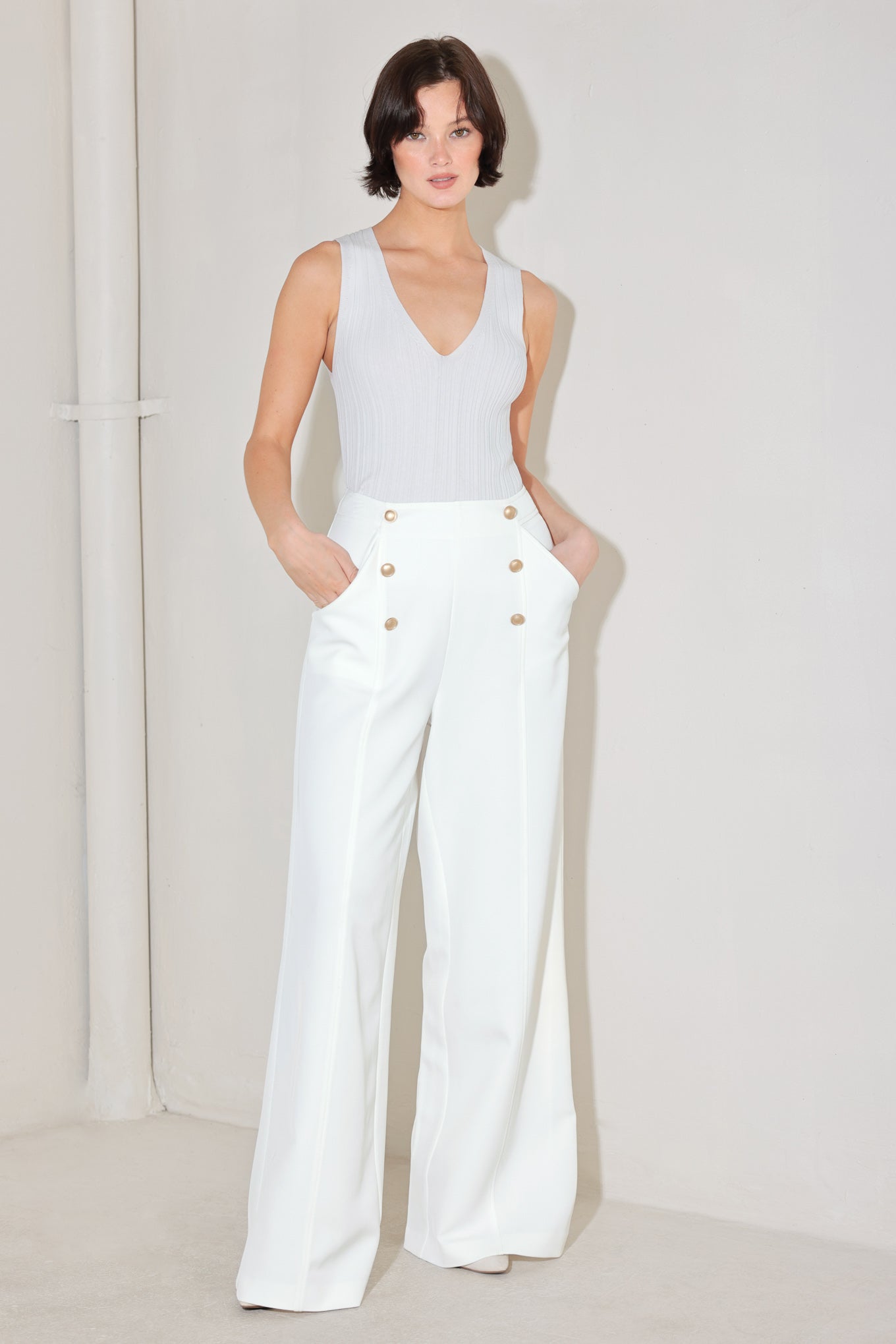 Woman wearing a white sleeveless top and wide-leg pants with gold buttons on a plain background