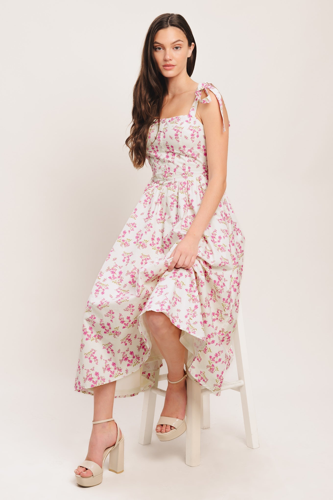 Woman wearing a floral dress sitting on a white stool against a plain background